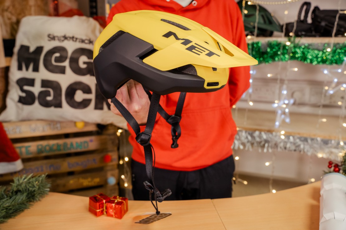 Close-up view of a MET Terrae MIPS helmet in bright yellow and black, held by a person against a festive background with decorations and a 'Mega Sack' sign.
